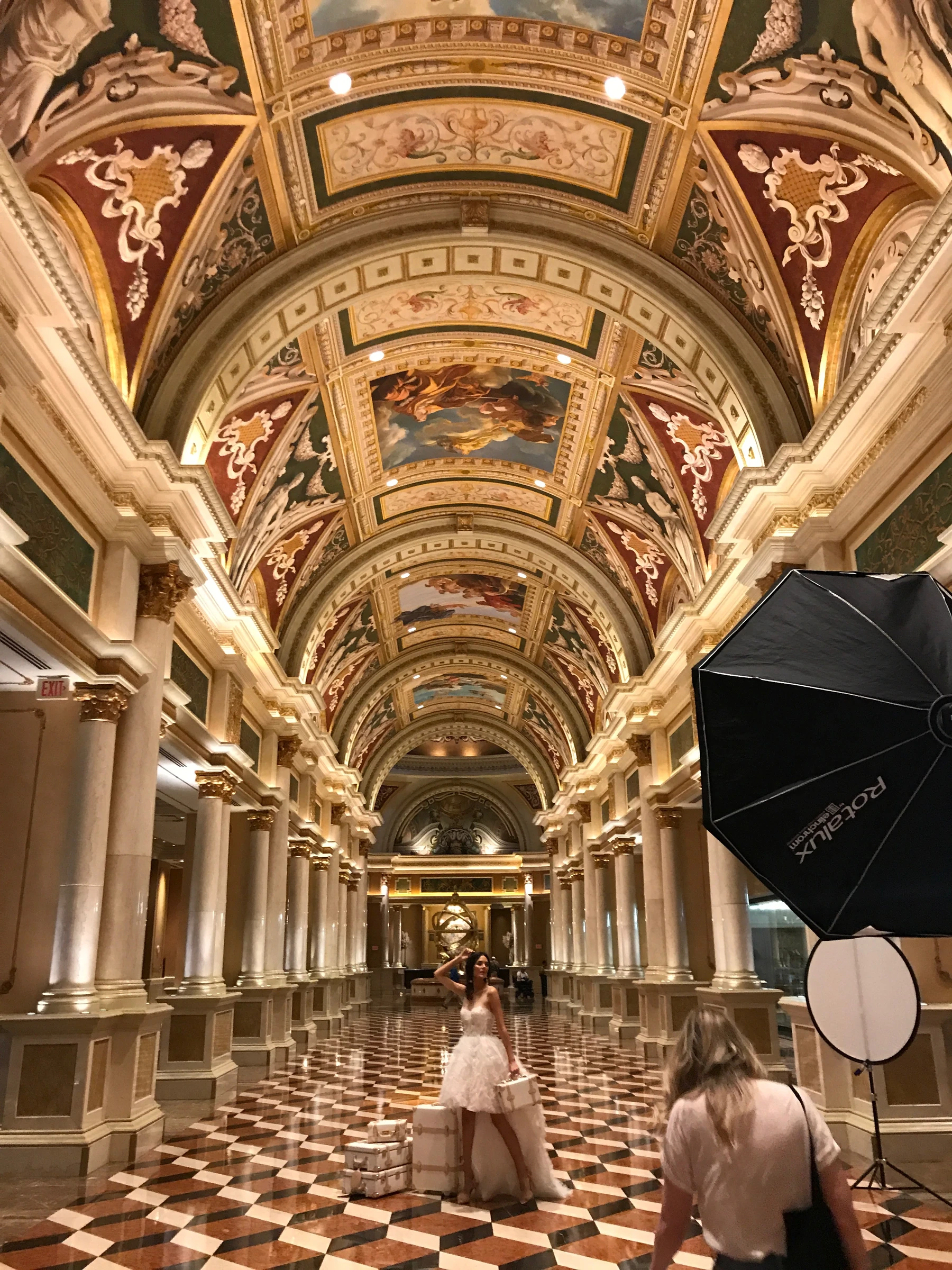 Behind-the-scenes location shoot in an ornate vaulted hall, with a subject posing in a white dress beside stacked suitcases, a softbox and reflector at right