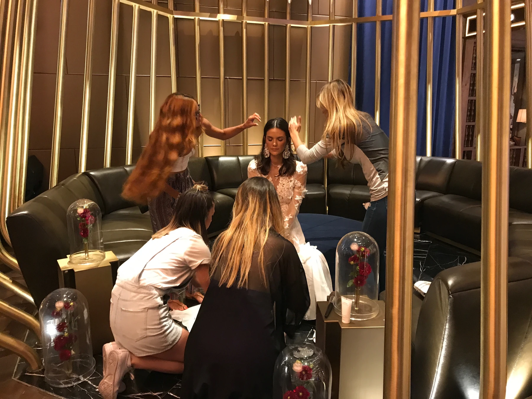 Behind the scenes, a team prepares a seated model in a floral-appliqué gown, adjusting hair and working around the skirt in a gold-accented lounge